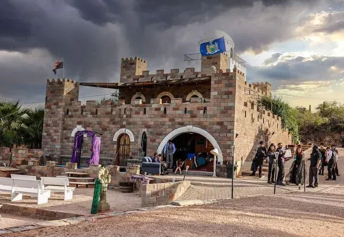 Stone castle-like building; people gather outside. Cloudy sky. Flag on tower, arched doorways.