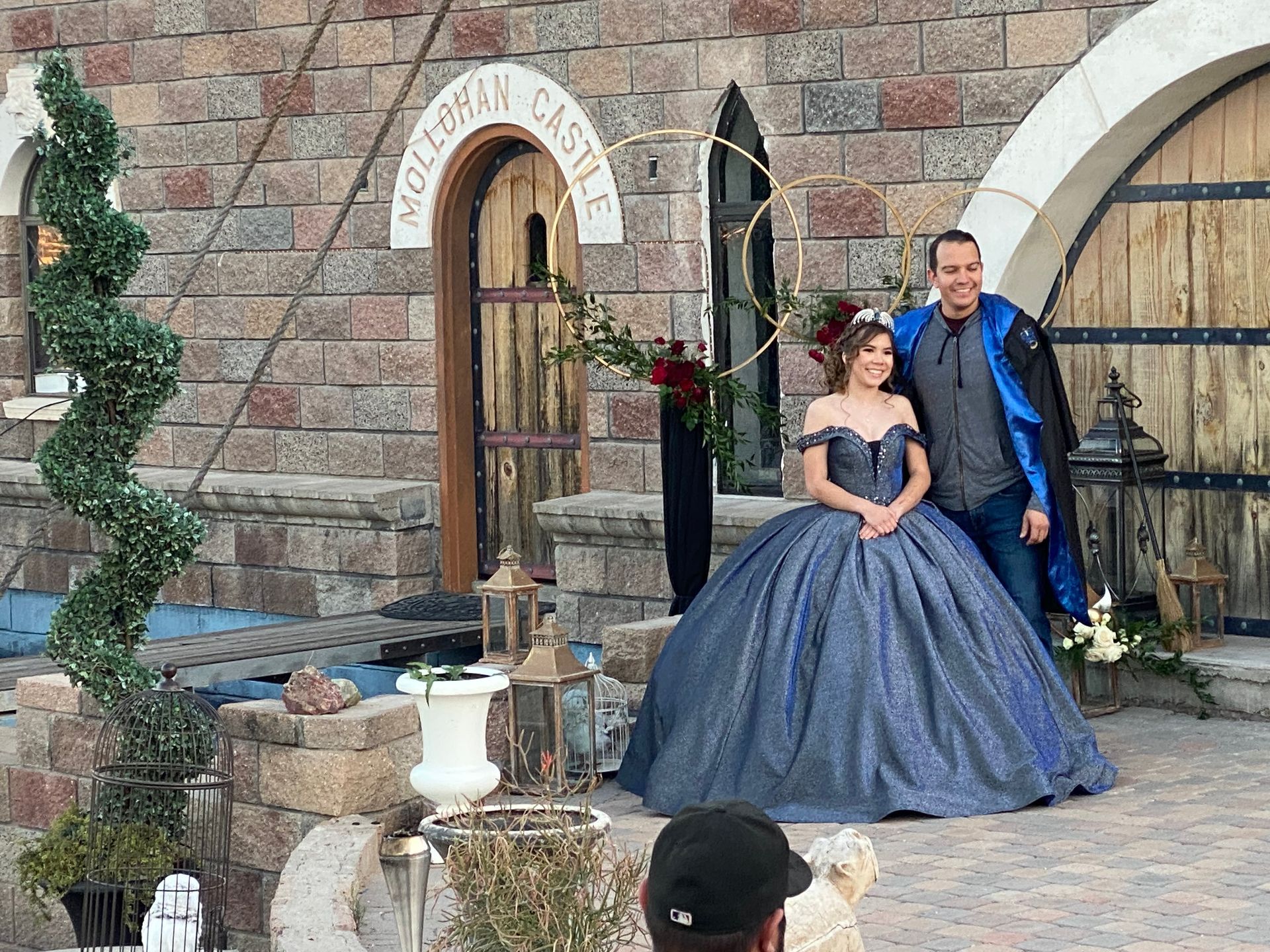 Couple posing for photo in front of a castle. Woman in blue ballgown, man in coat, building brick facade.