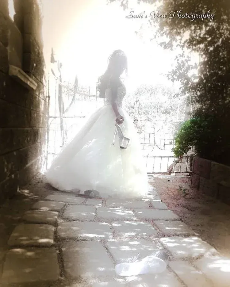 Bride in white gown holding shoes, standing in sunlight by stone path.