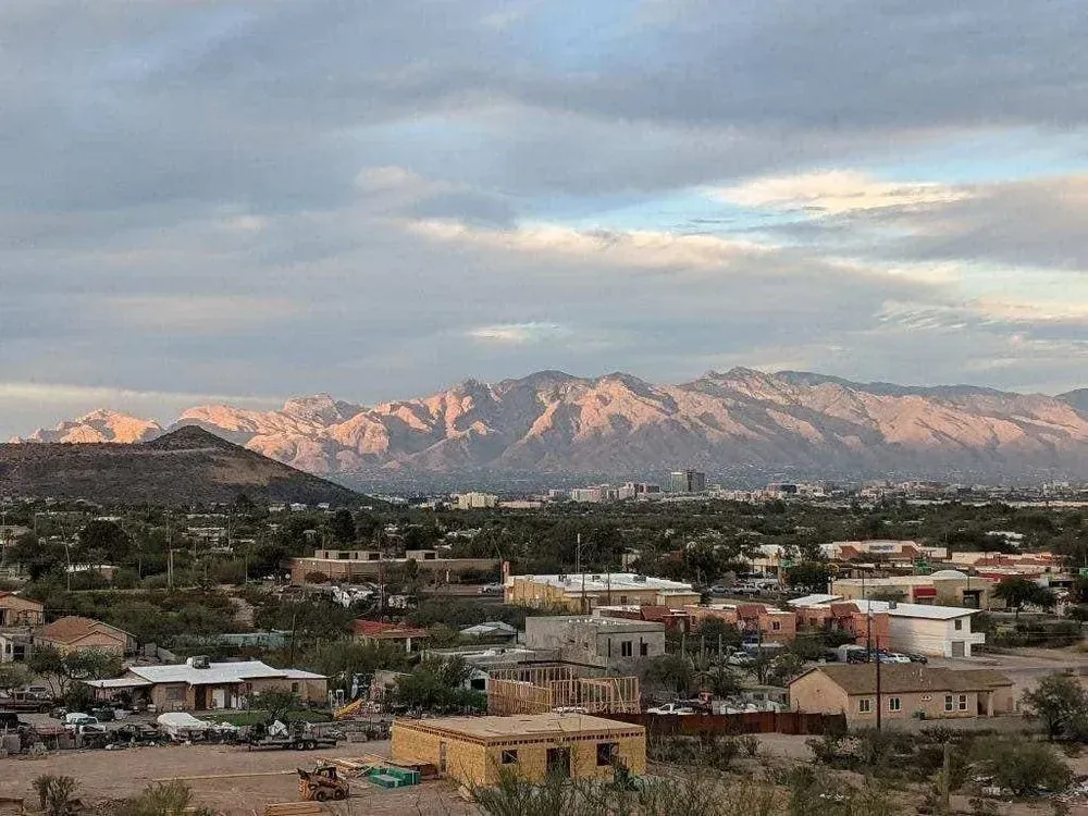 Houses and buildings in a desert landscape with mountains under a cloudy sky.