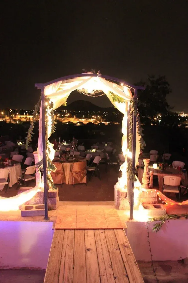 Wooden archway lit with lights, leading to a wedding reception at night. Tables set, city lights in background.