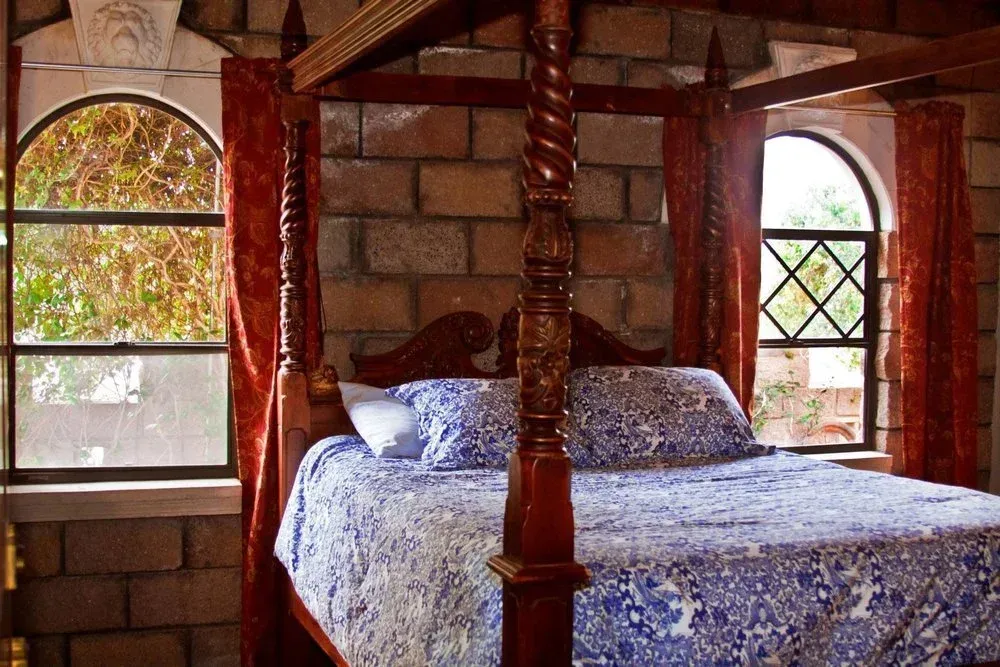 Bedroom with stone walls, four-poster bed with blue floral quilt. Two arched windows with red curtains.