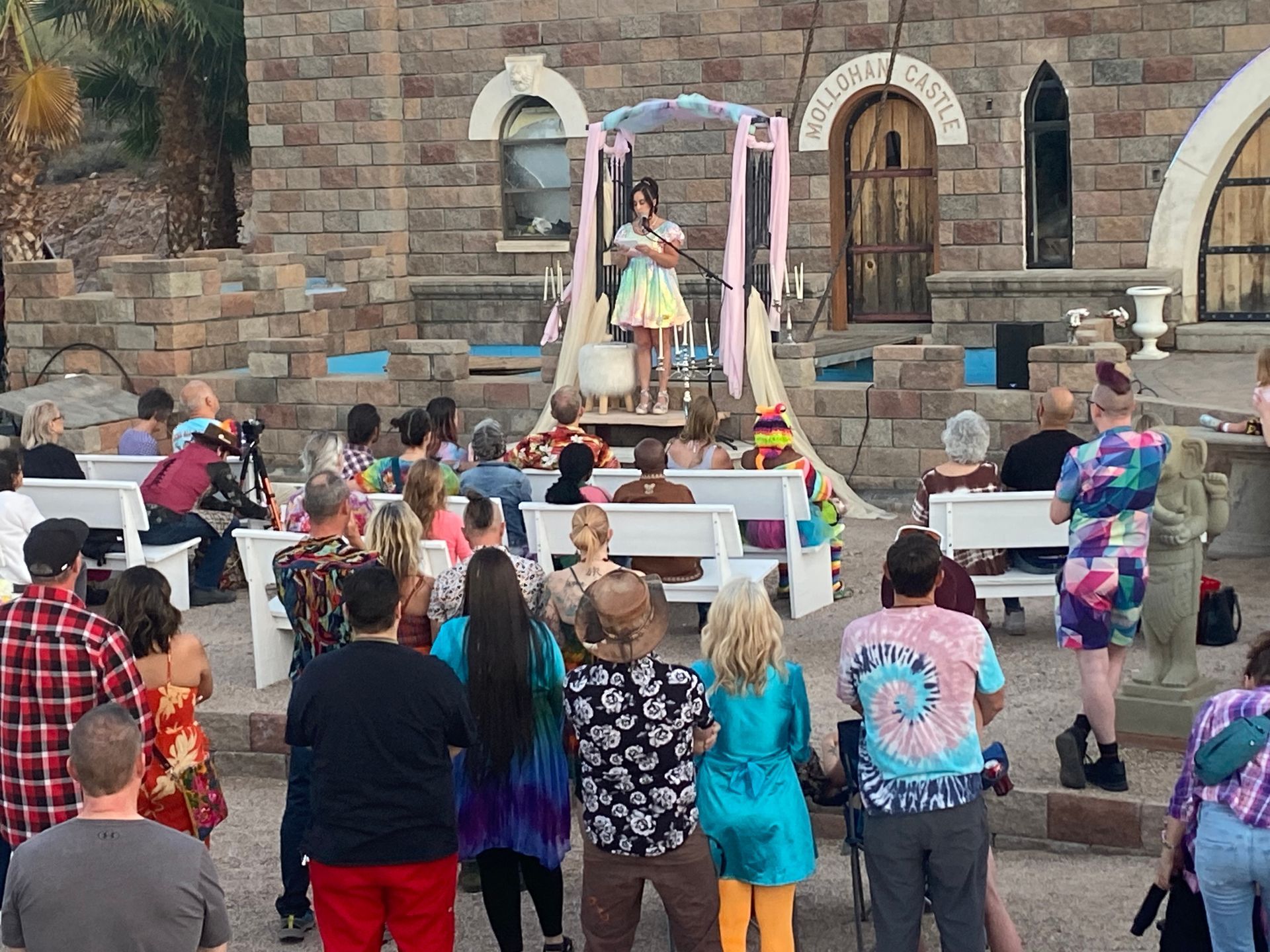 A person plays music on a stage with an audience outdoors in front of a stone building with an arched doorway.