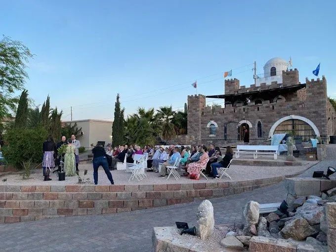 Outdoor event at a stone castle-like building. People seated in chairs, some standing, with blue sky backdrop.
