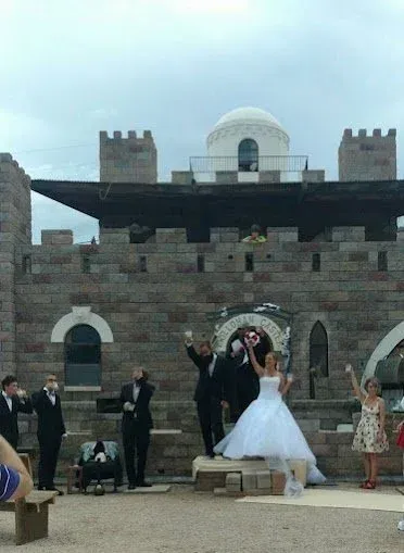 Wedding party toasting outside a stone castle; bride in white gown, guests in formal attire. Cloudy sky.
