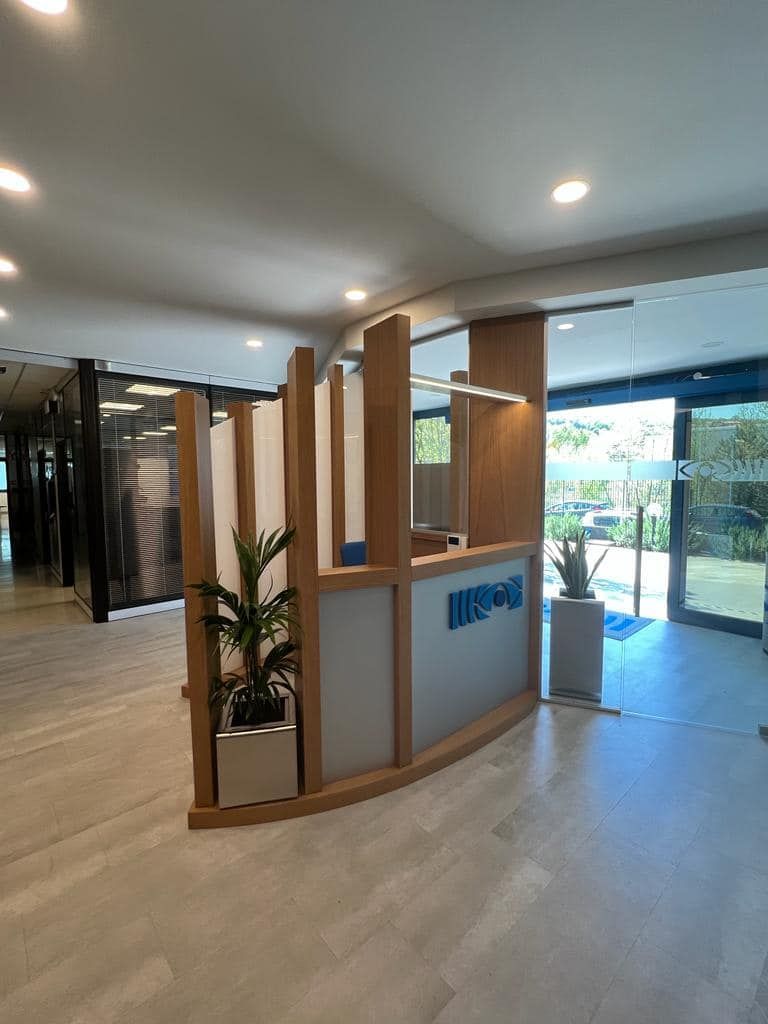 Hall moderna con bancone della reception in legno curvato, porte in vetro, pianta in vaso e luminosa luce naturale.
