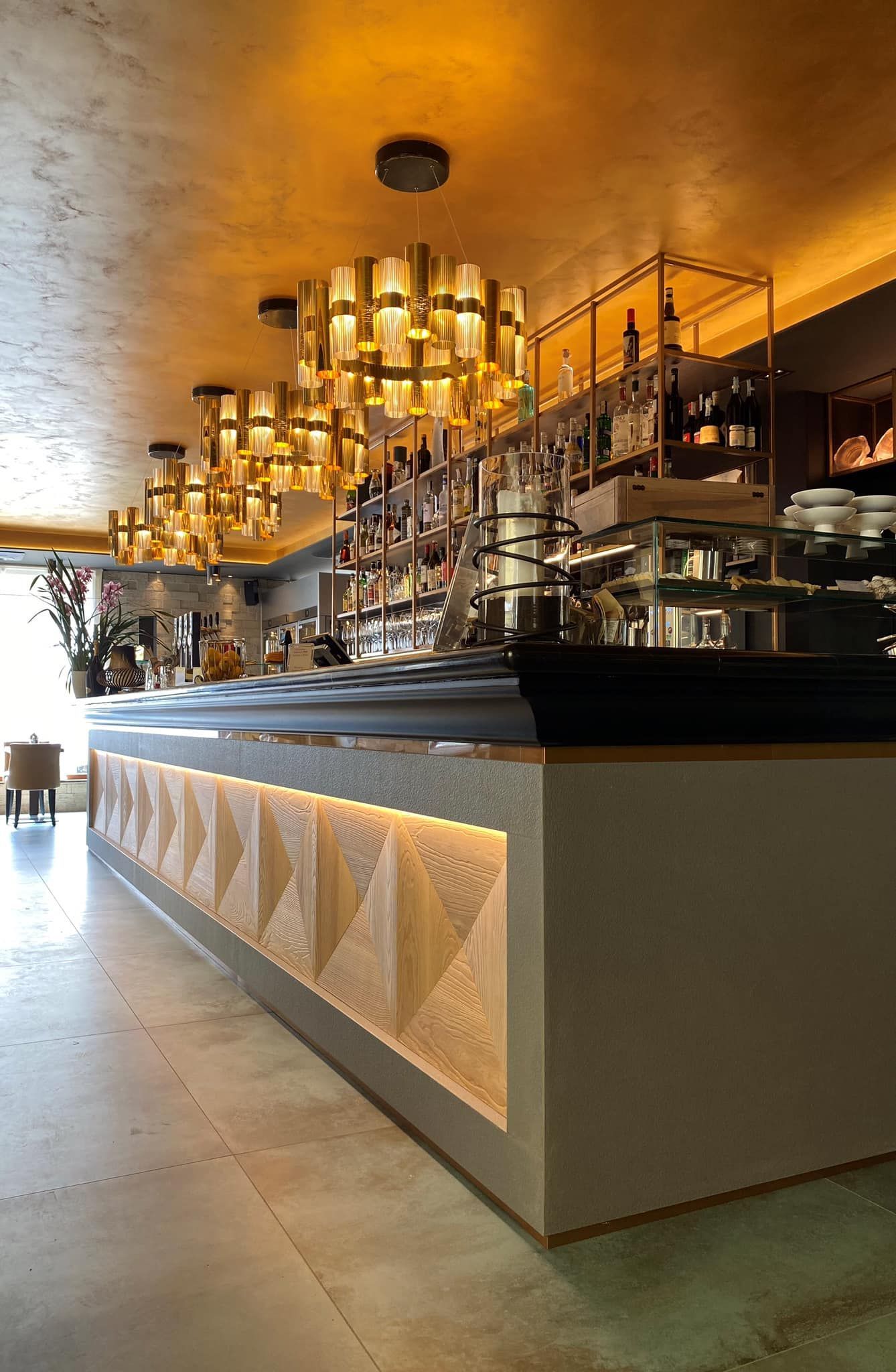 Interno di un bar moderno e accogliente, con un lungo bancone, lampade a sospensione dorate e scaffali pieni di bicchieri e bottiglie.