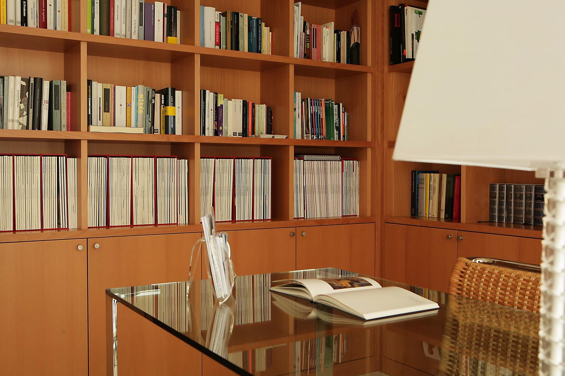 Studio con pannelli in legno, librerie, scrivania in vetro e lampada da tavolo in primo piano.