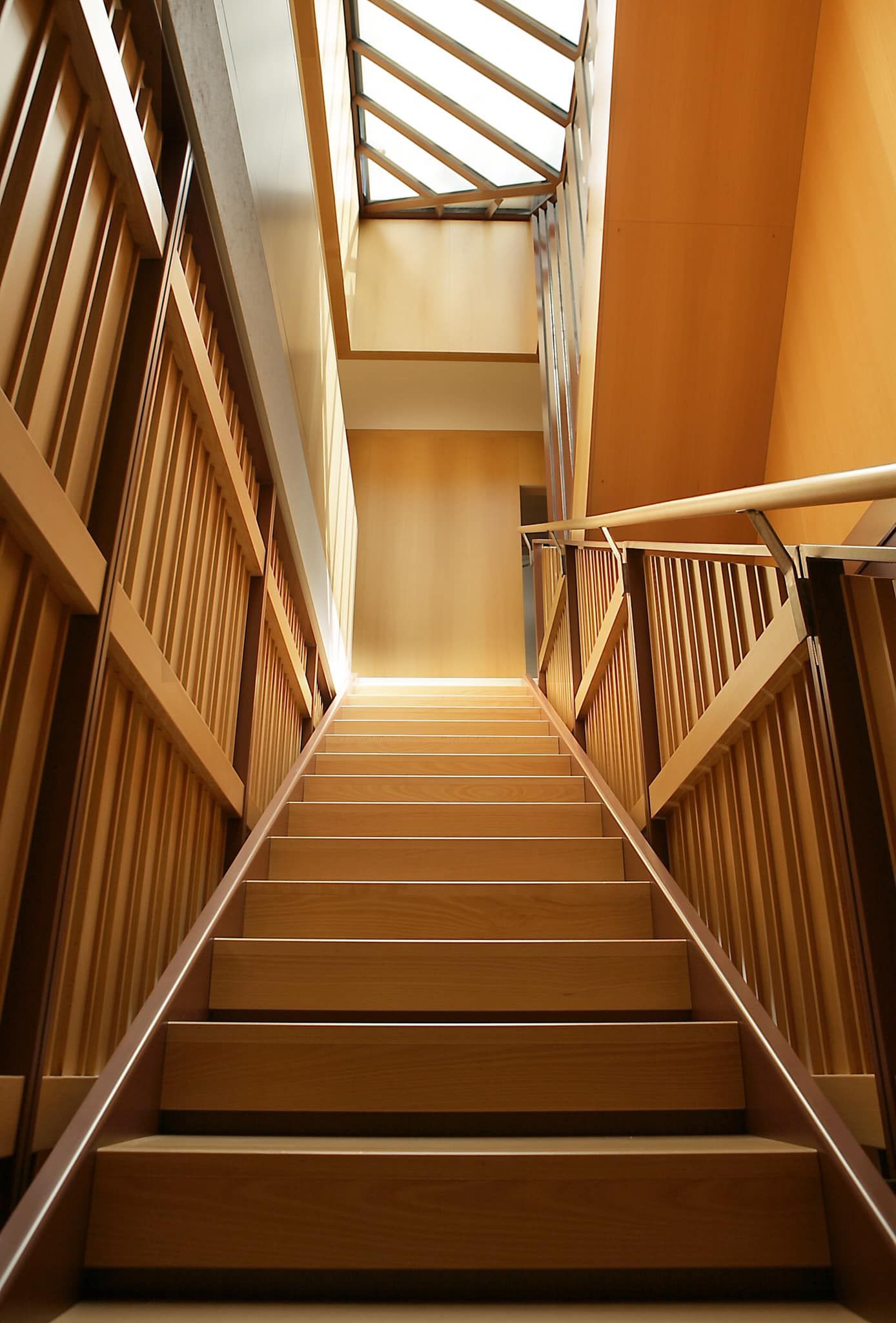 Scala in legno in un interno illuminato da una luce calda, con ringhiere a traliccio e un lucernario sopra.