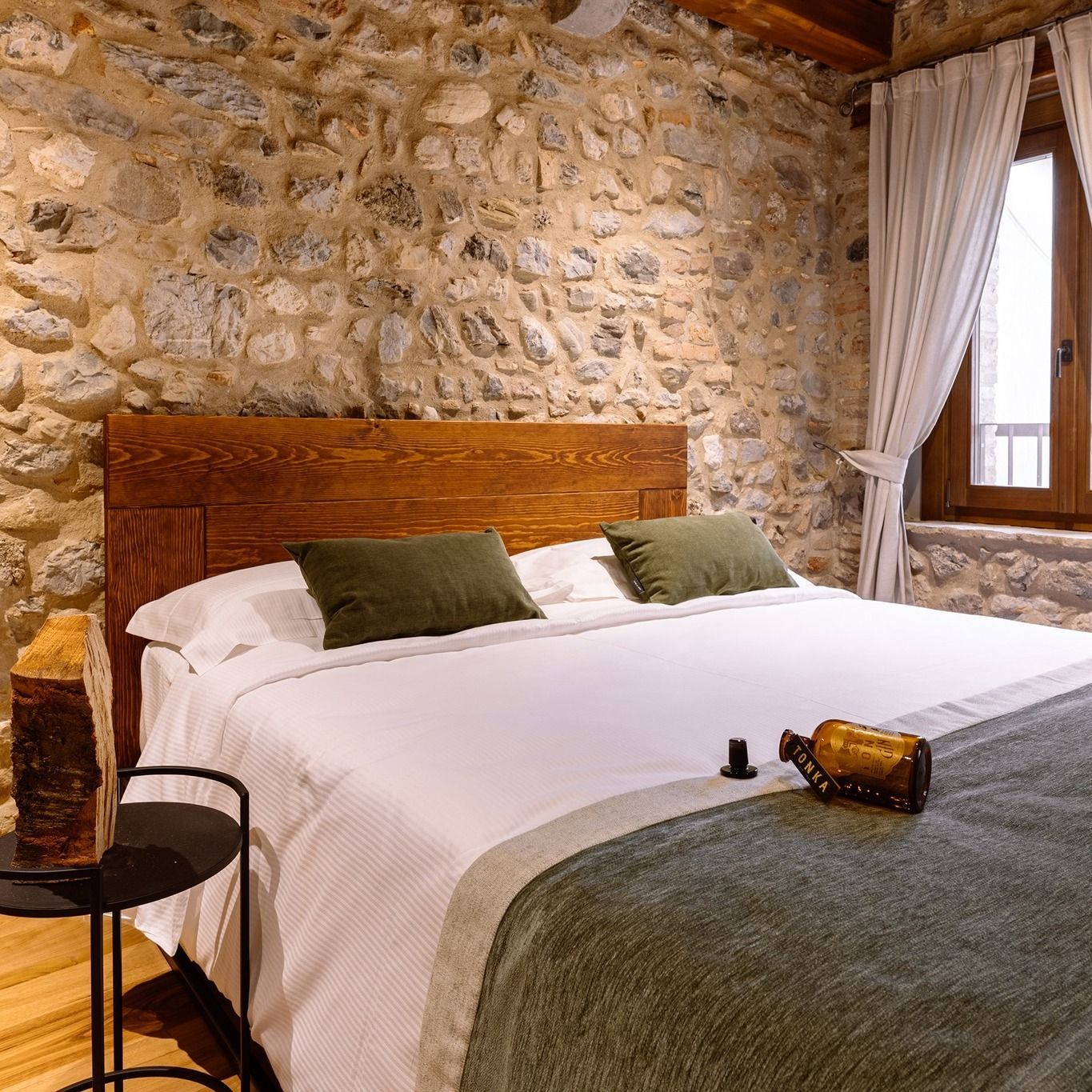 Camera da letto con pareti in pietra, letto in legno, lenzuola bianche, coperta verde e finestra con tende.