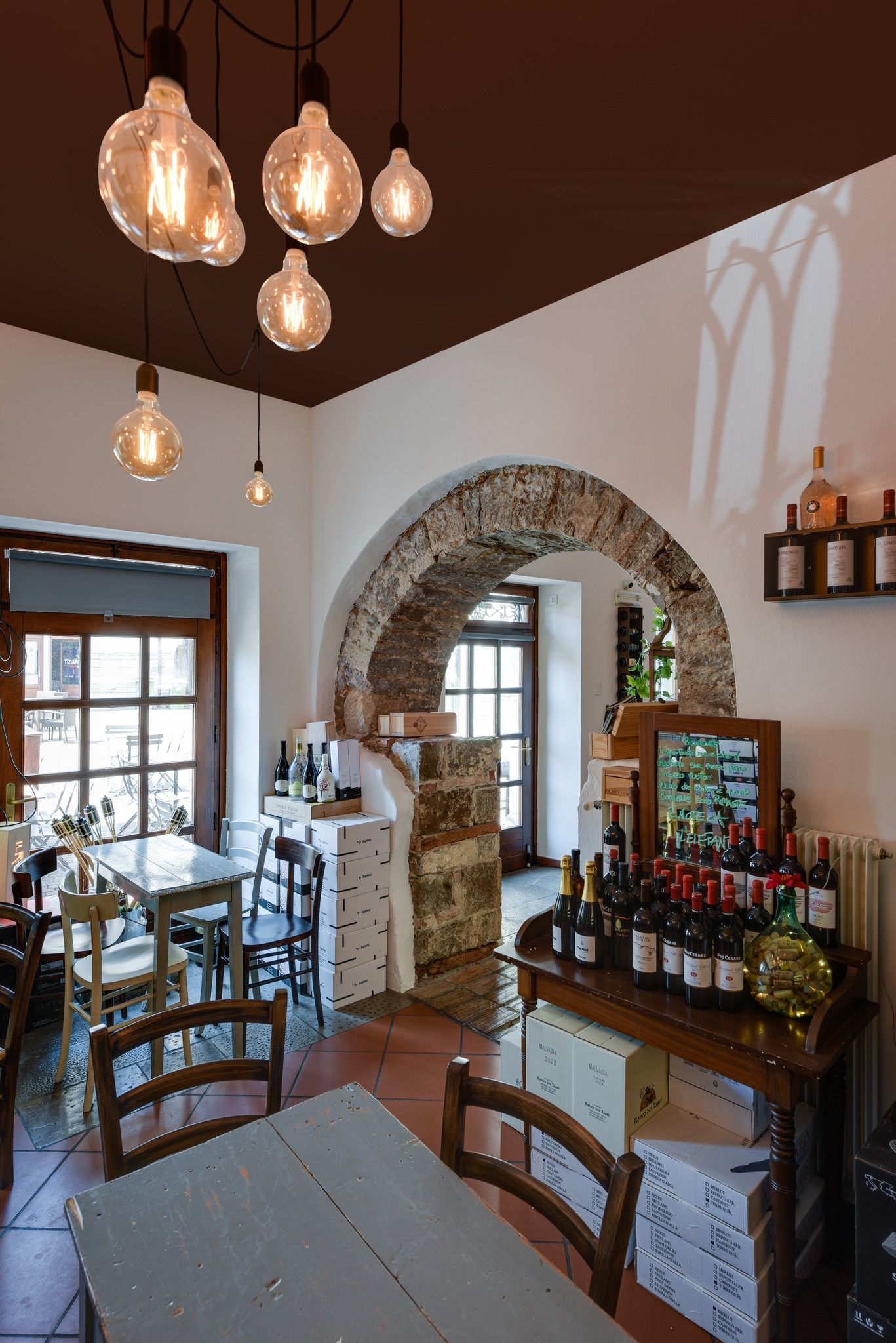 Accogliente interno di un'enoteca con tavoli rustici, lampade a sospensione e porta ad arco in pietra.