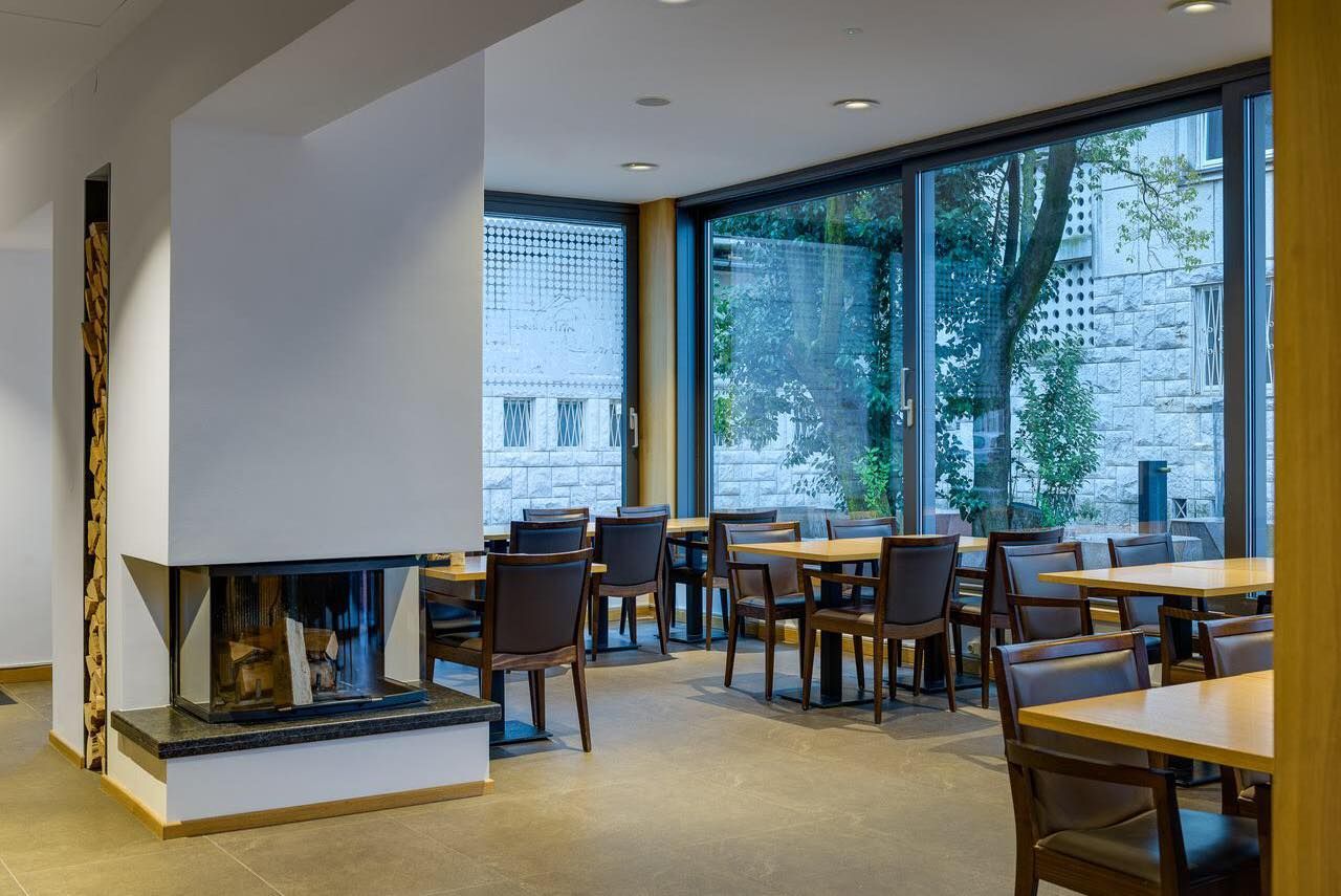 Sala da pranzo moderna con tavoli, sedie, camino e ampie finestre che si affacciano su alberi ed edifici.
