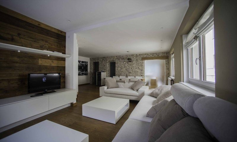 Soggiorno moderno con divano angolare bianco, parete con inserti in legno, TV e ampie finestre con tende.