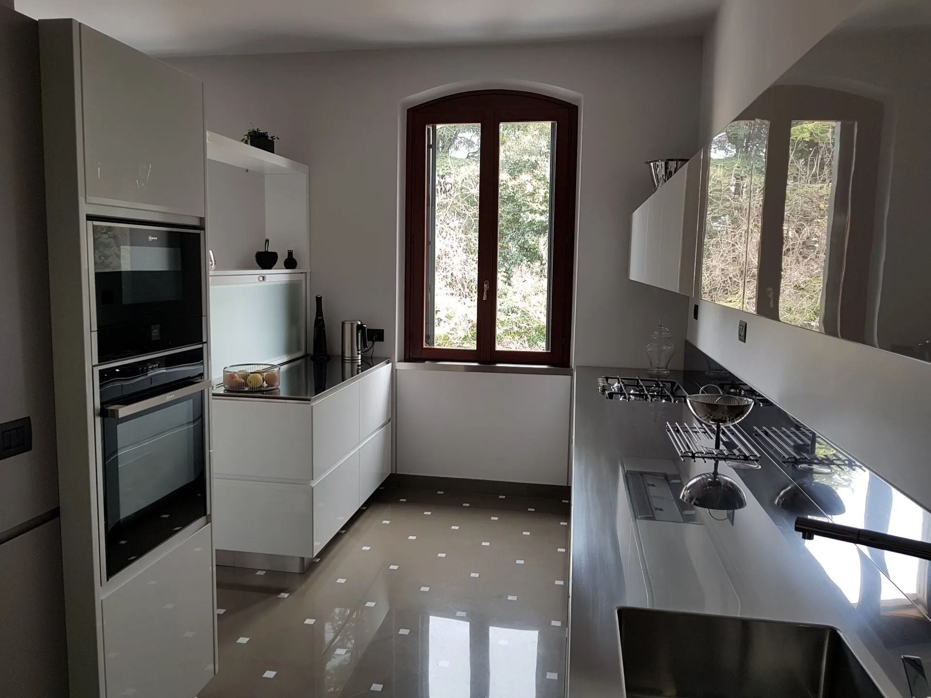 Cucina moderna con mobili bianchi, forni a incasso e lunghi banconi accanto a una finestra.