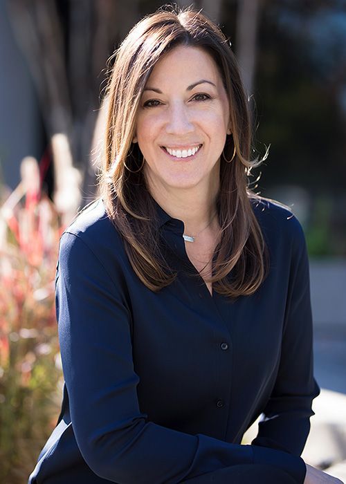 Smiling woman in a navy blouse seated outdoors with blurred plants in the background