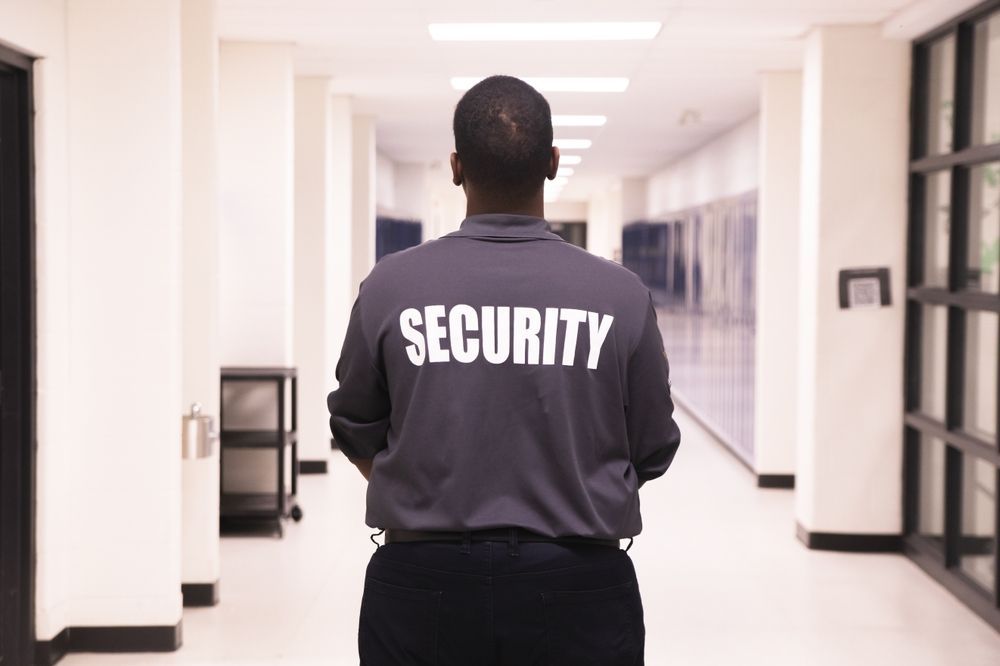 Security guard walking in a bright hallway, seen from behind, wearing a jacket labeled “SECURITY”