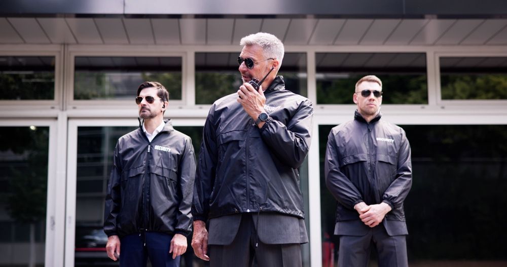 Three men in dark jackets stand outside a building, with the center man holding a hand to his chin.