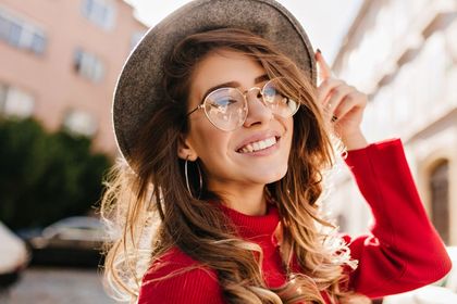 Woman with hat and glasses smiles, pointing upward outdoors. Red sweater, city background.