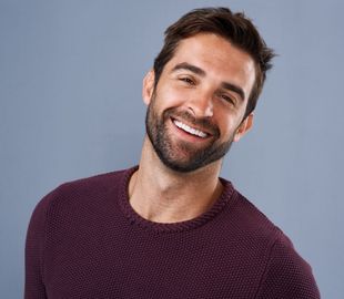Man with brown hair and beard smiles, wearing a purple sweater, against a gray backdrop.