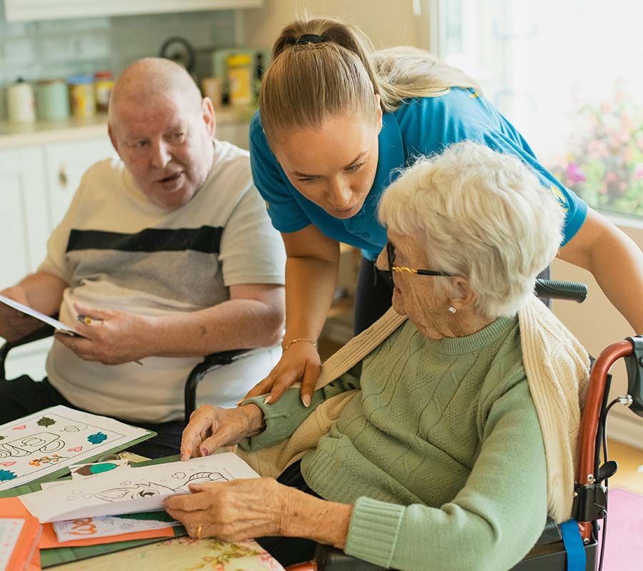 A young woman leans over elderly people in wheelchairs, looking at artwork, indoors.