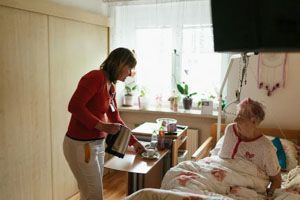 Woman pouring liquid from a kettle into a cup for a person in bed in a room with a window.