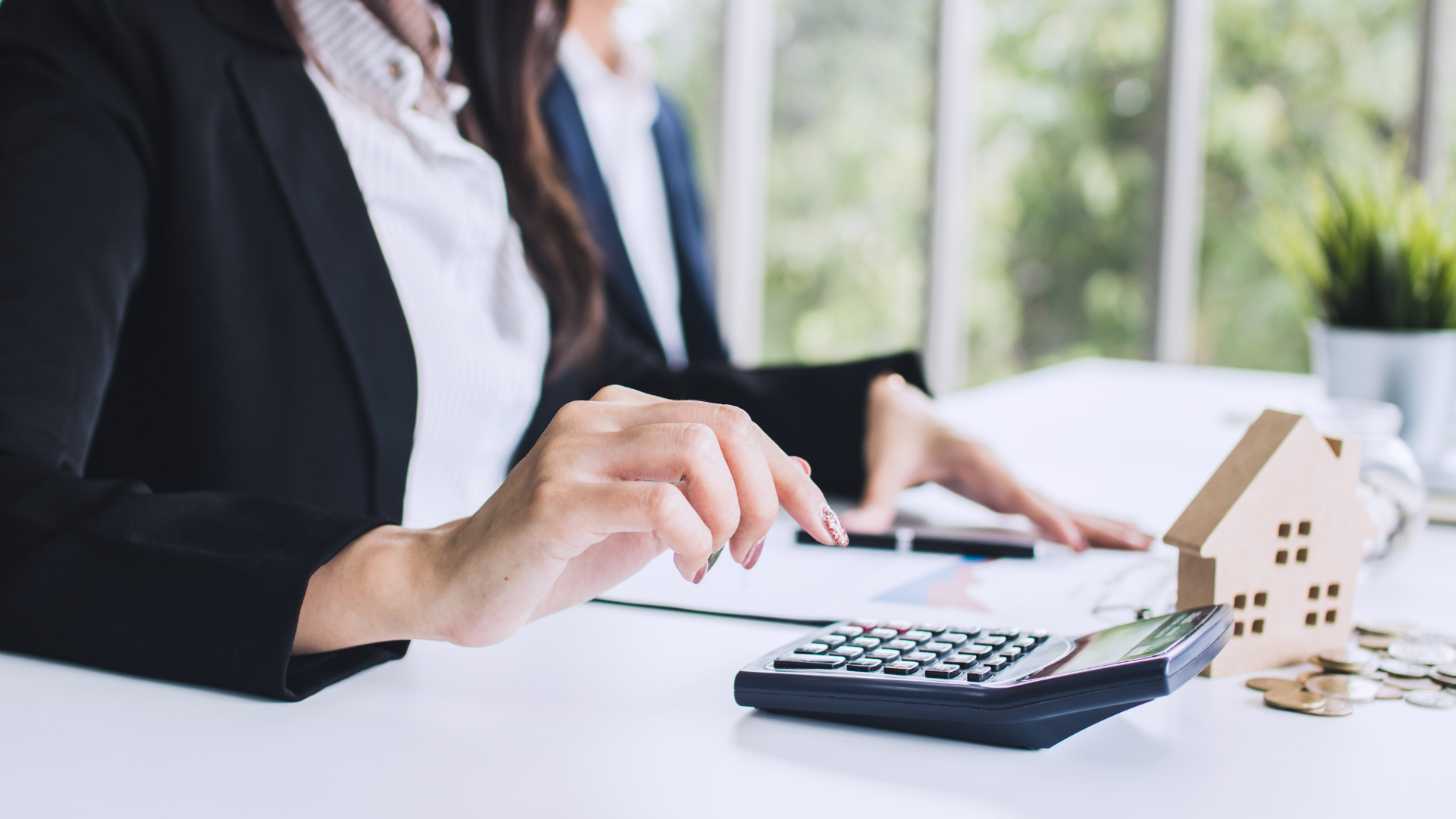 A person in business attire uses a calculator next to a small wooden house model and coins on a white desk.