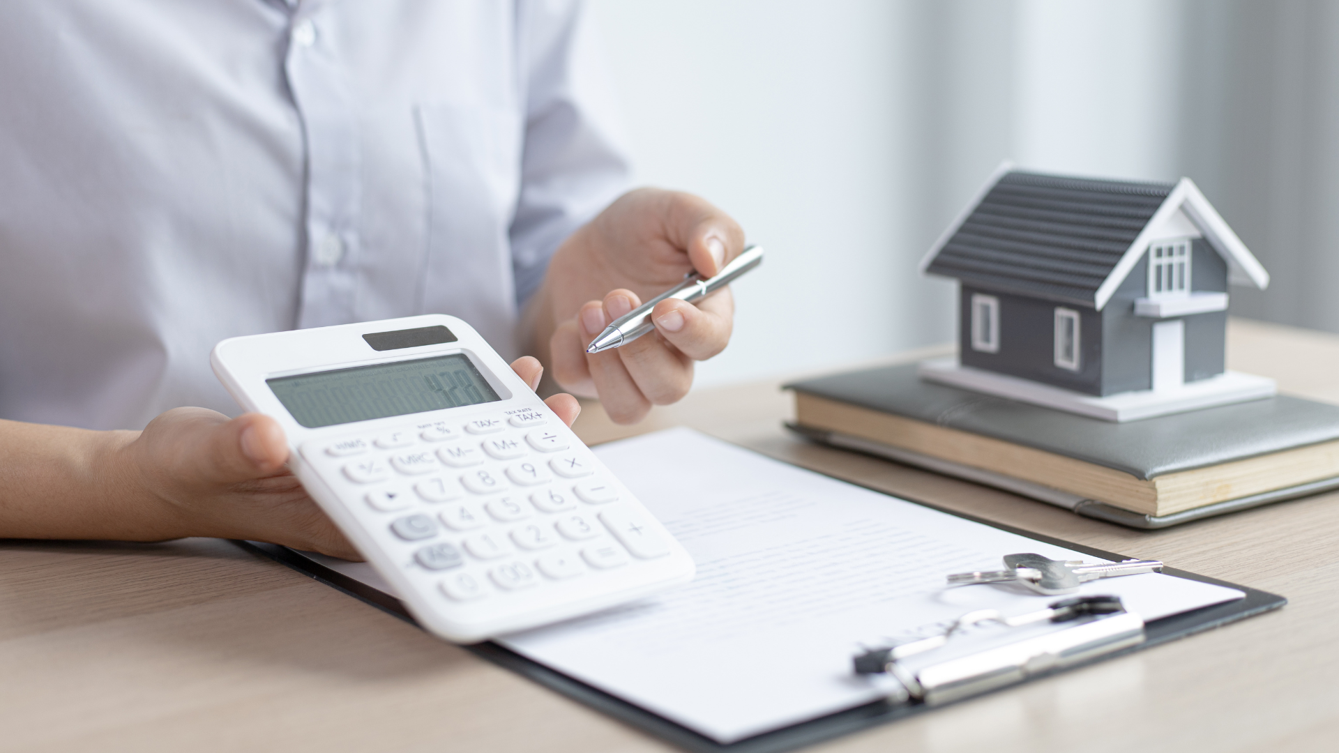 A person in a light shirt holds a calculator and pen next to a model house on a desk with a clipboard.