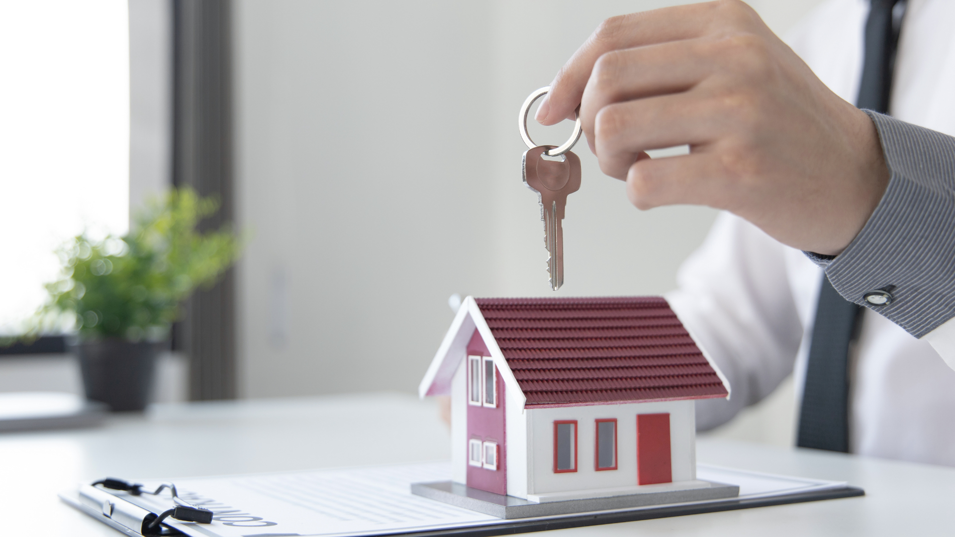 A person in professional attire holds a key above a model house sitting on a clipboard at a desk.