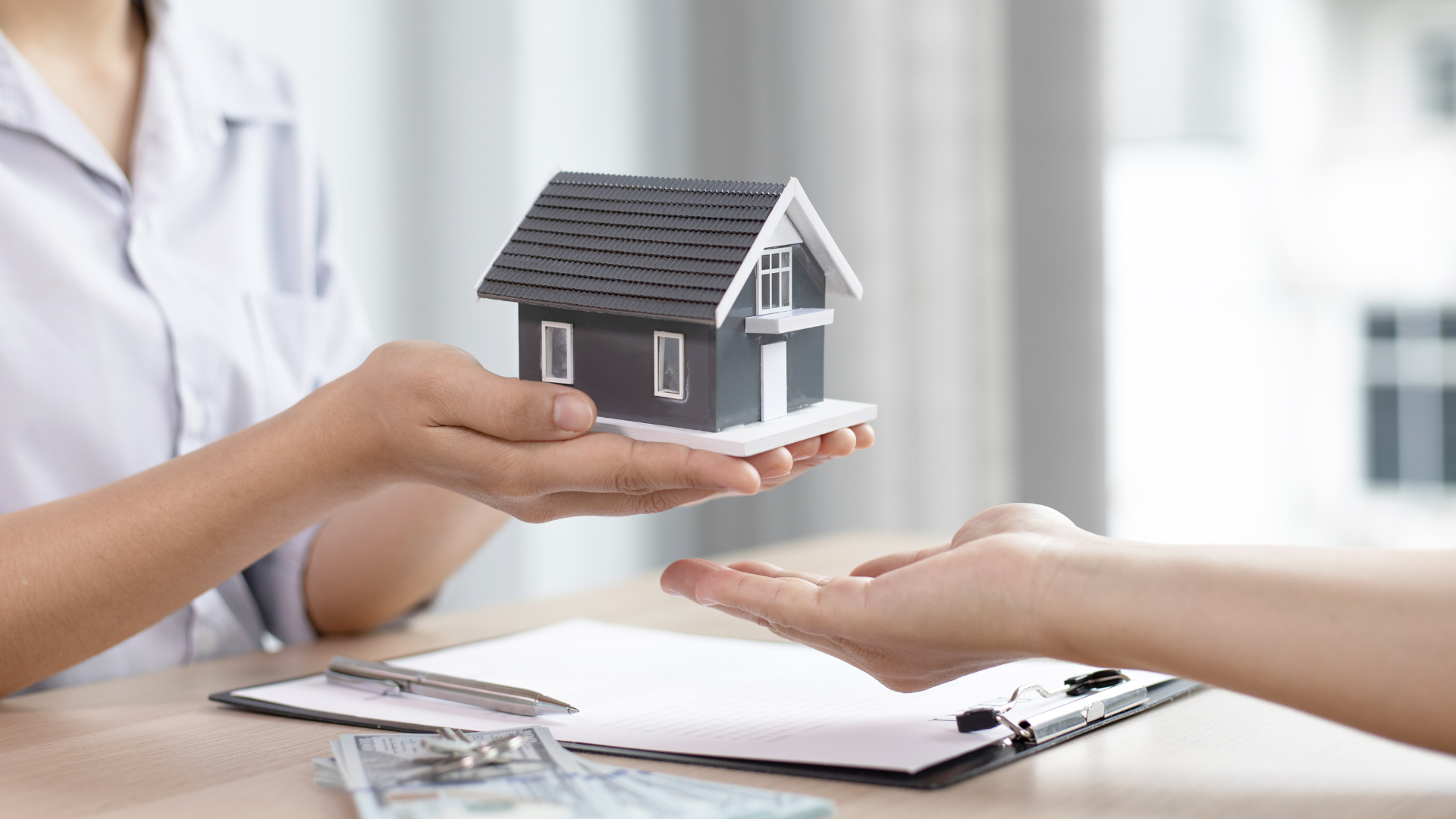 One person hands a model house to another person over a desk with paperwork and money.