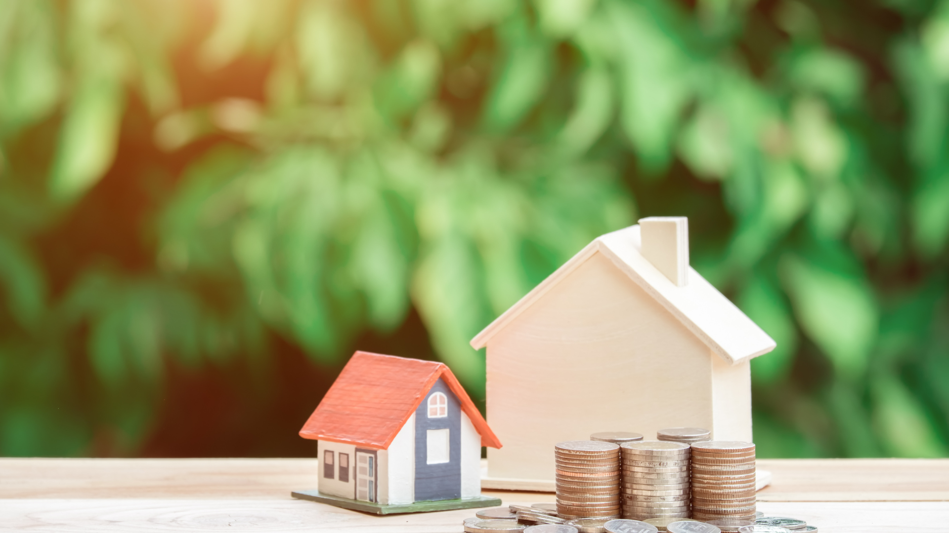 Two toy houses and stacks of coins on a table against a blurred green background.