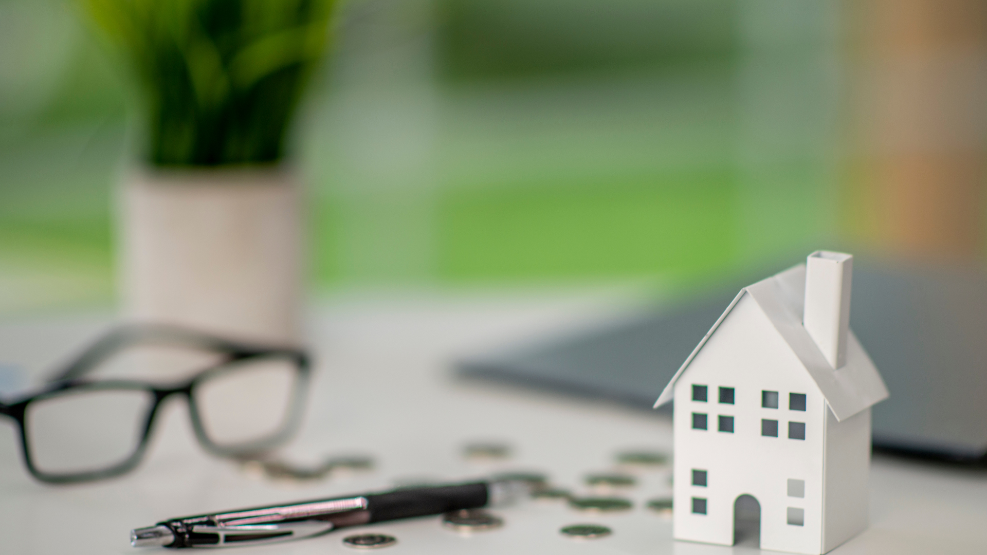 A white model house sits on a desk with a pen, eyeglasses, and scattered coins, symbolizing home finance and real estate.