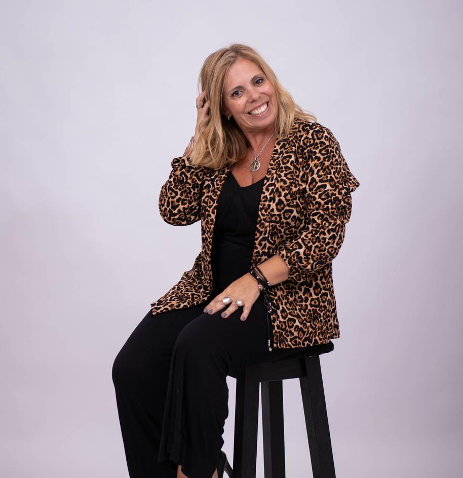 Mujer con chaqueta con estampado de leopardo, mono negro, sentada en un taburete, sonriendo, con la mano en el cabello.