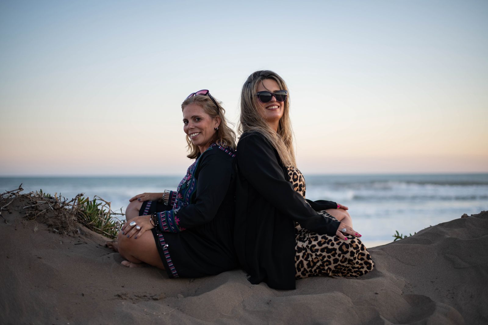 Dos mujeres sentadas espalda con espalda en una duna de arena, con el océano de fondo. Colores del atardecer, sonrisas.