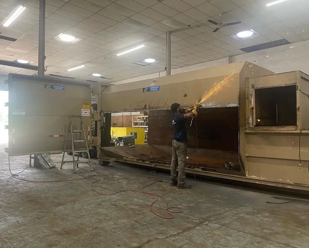 A person using a grinder to cut metal on a large, tan metal structure inside a workshop.