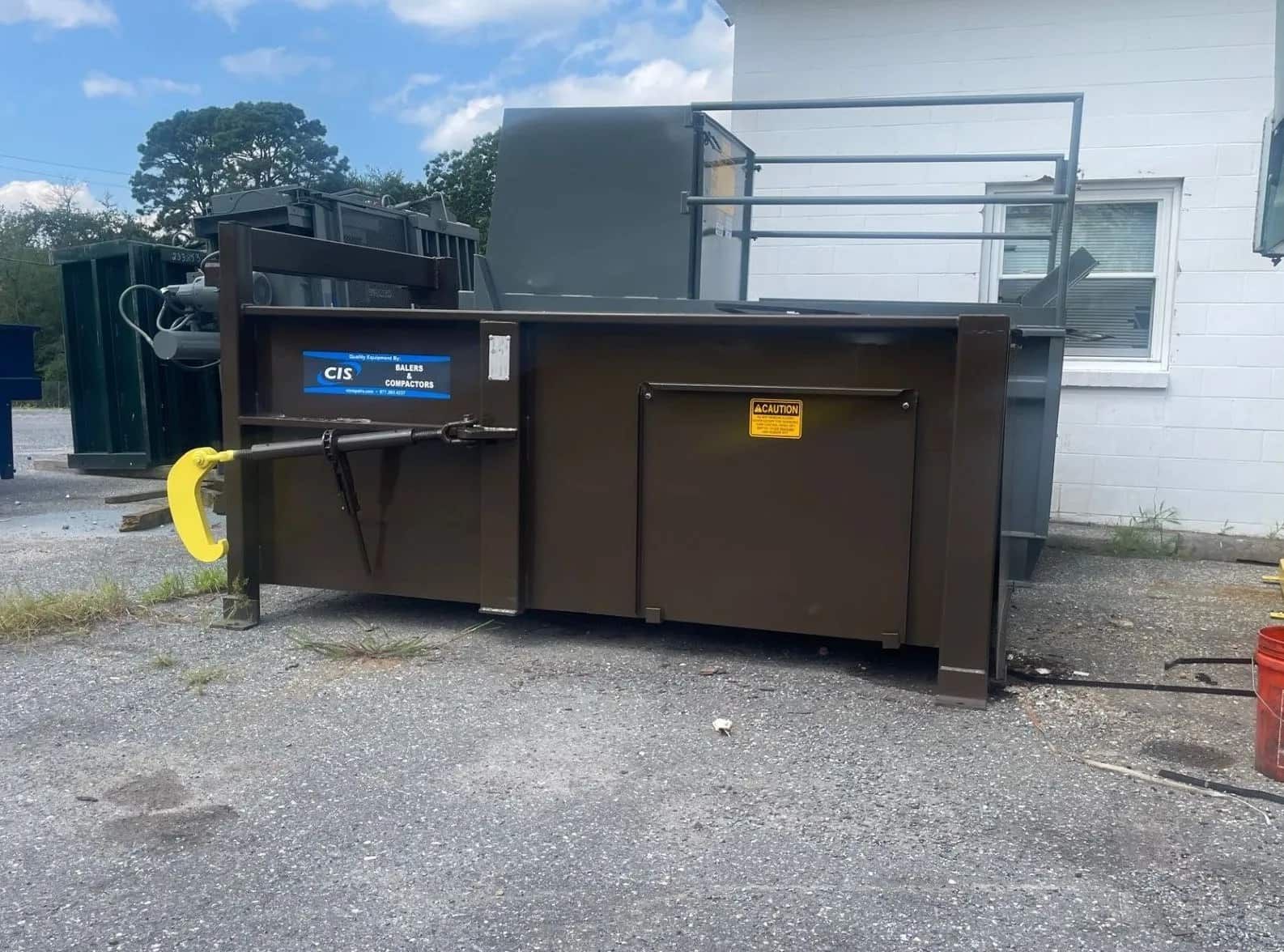 Brown industrial trash compactor on a concrete surface, beside a building.