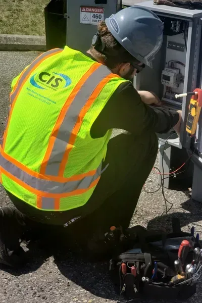 Worker in safety vest and hard hat working on electrical equipment outdoors.