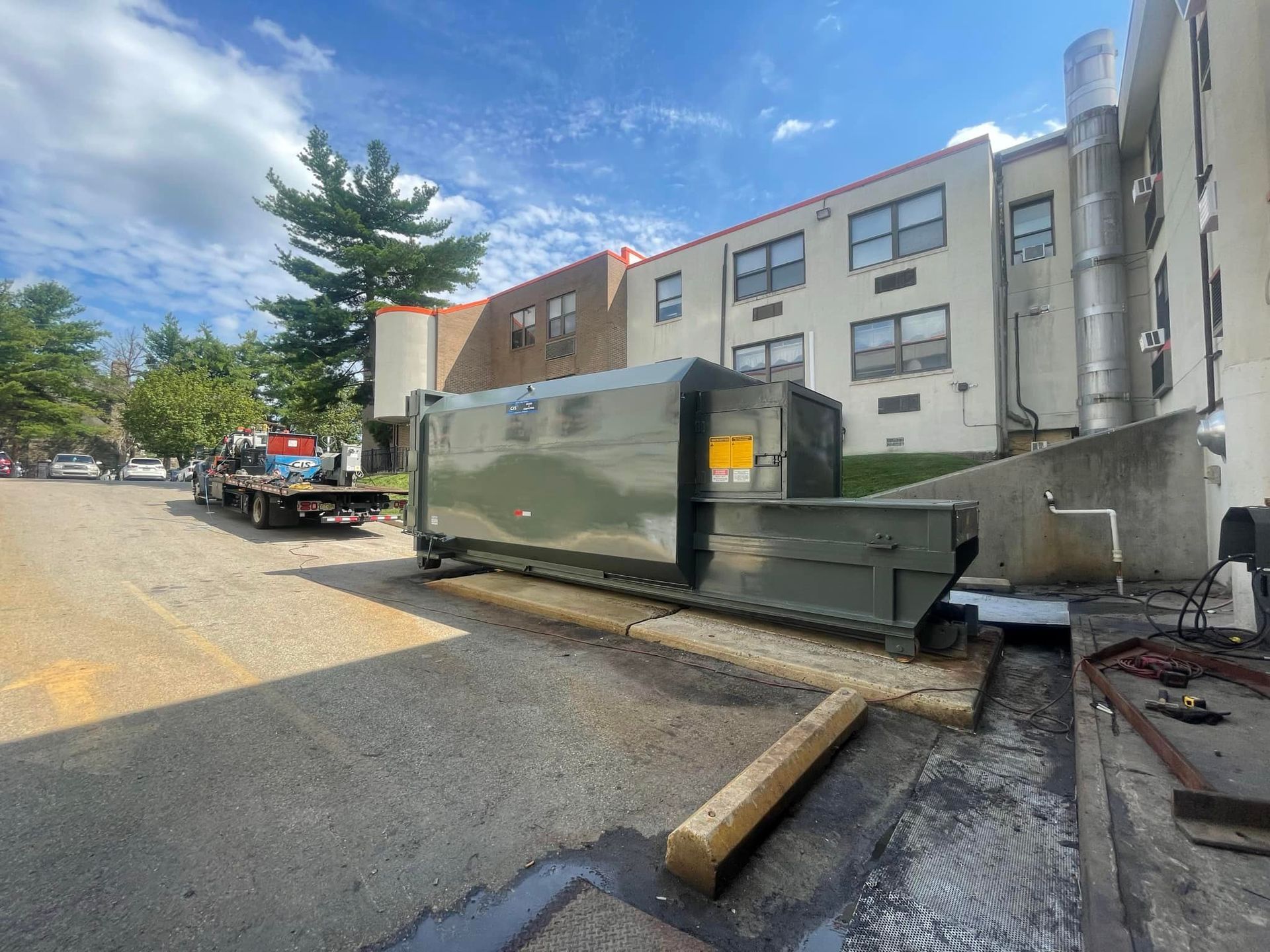 Large, olive green dumpster being serviced by a flatbed truck in a parking area near a building.