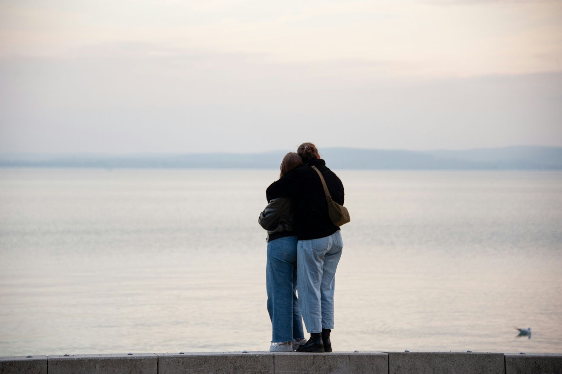 Two people hugging, overlooking a calm body of water at dusk.