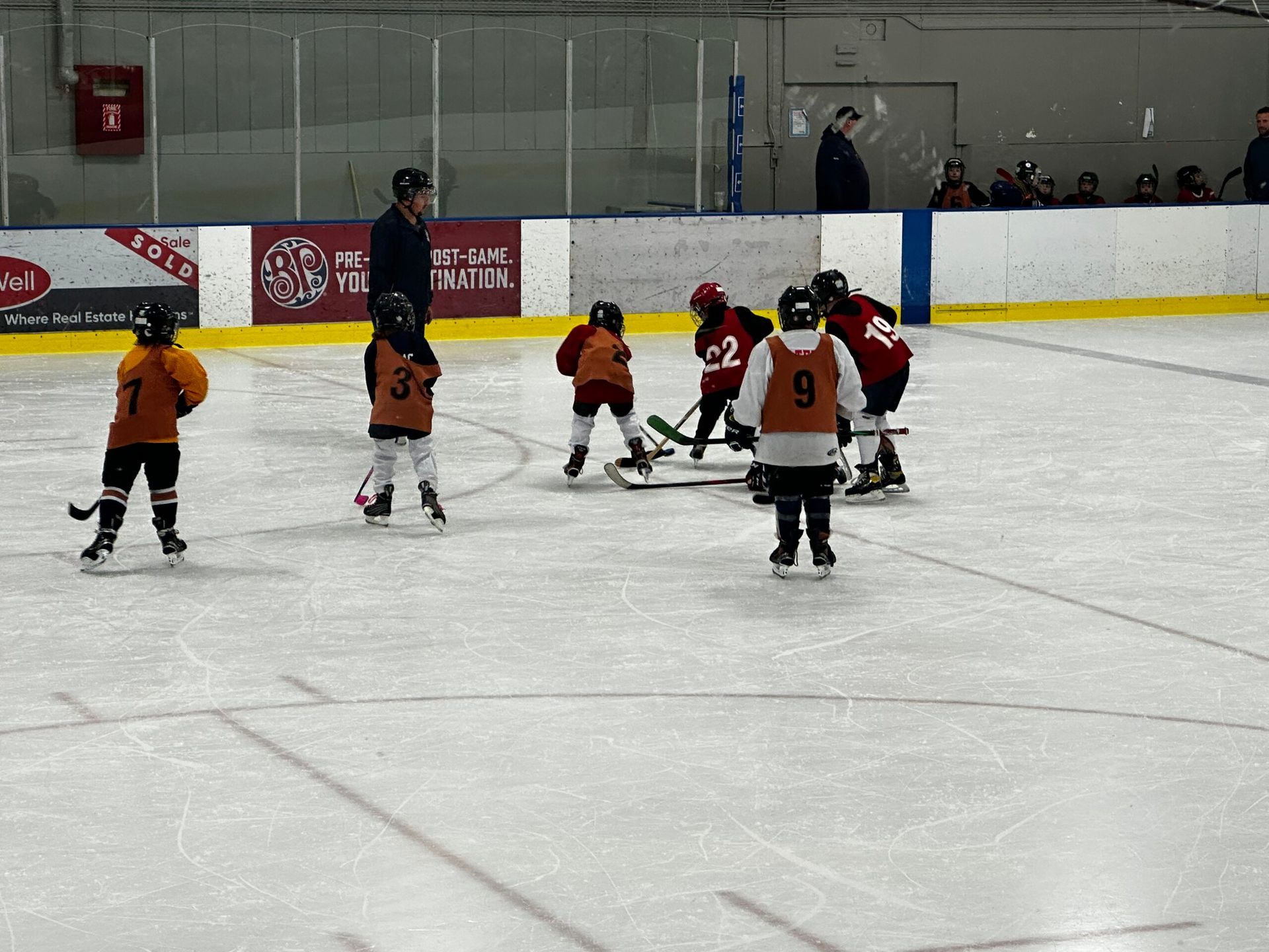 Children in orange and red jerseys play hockey on an indoor ice rink.