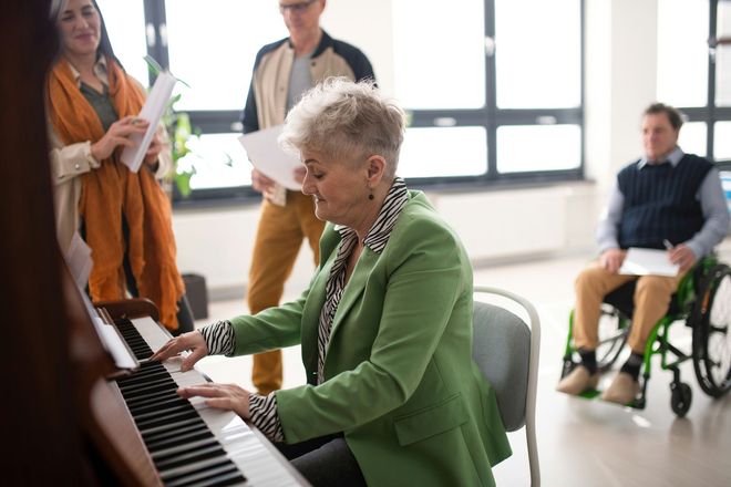 Woman playing piano in front of a group holding sheet music. A person in a wheelchair is also present.
