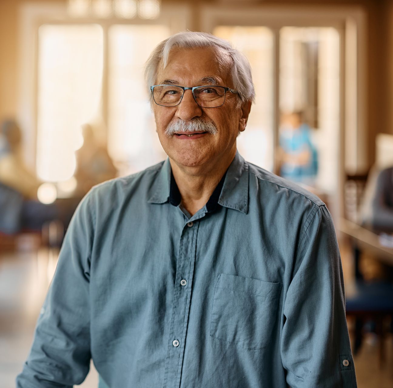 Smiling older man with glasses in a blue shirt, indoors with people blurred in the background.