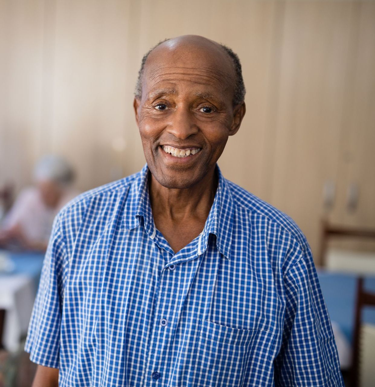 Smiling elderly Black man wearing a blue checked shirt, standing indoors.