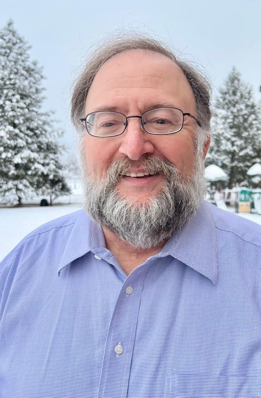 A man with a beard and glasses is smiling in front of a snowy forest.