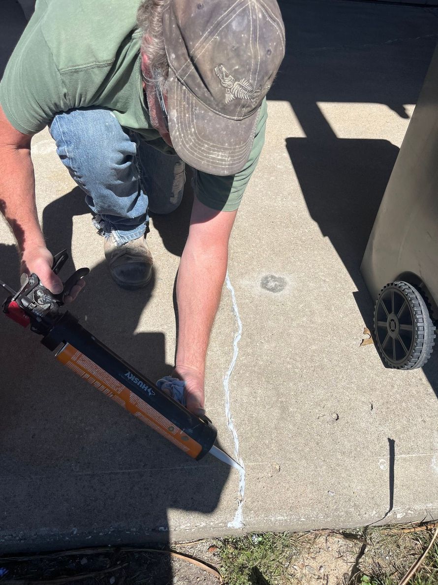 Person using a caulking gun to seal a crack in concrete beside a wheelbarrow.