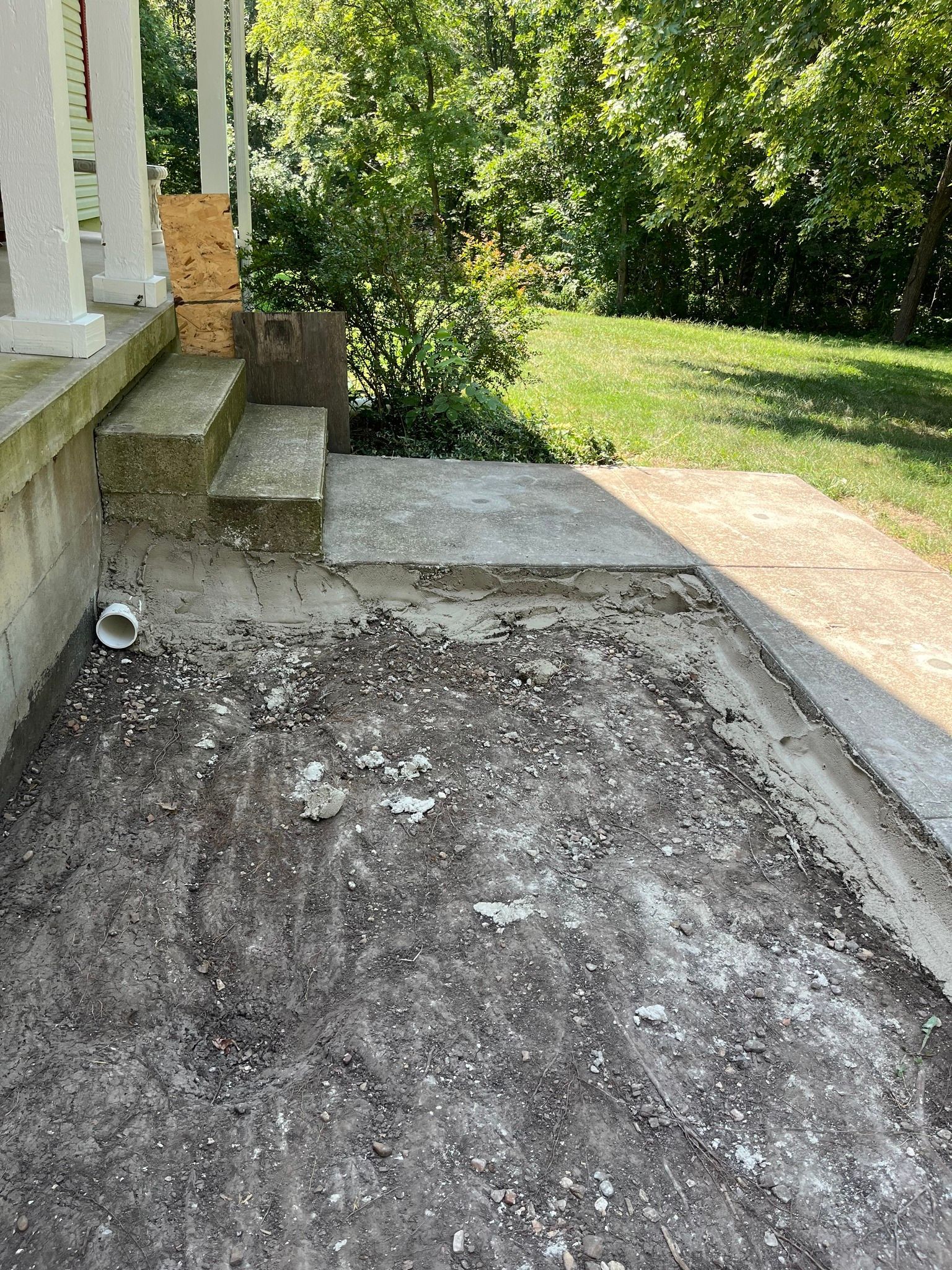 Rough gravel path beside house steps and concrete patio, leading to a grassy yard with trees.