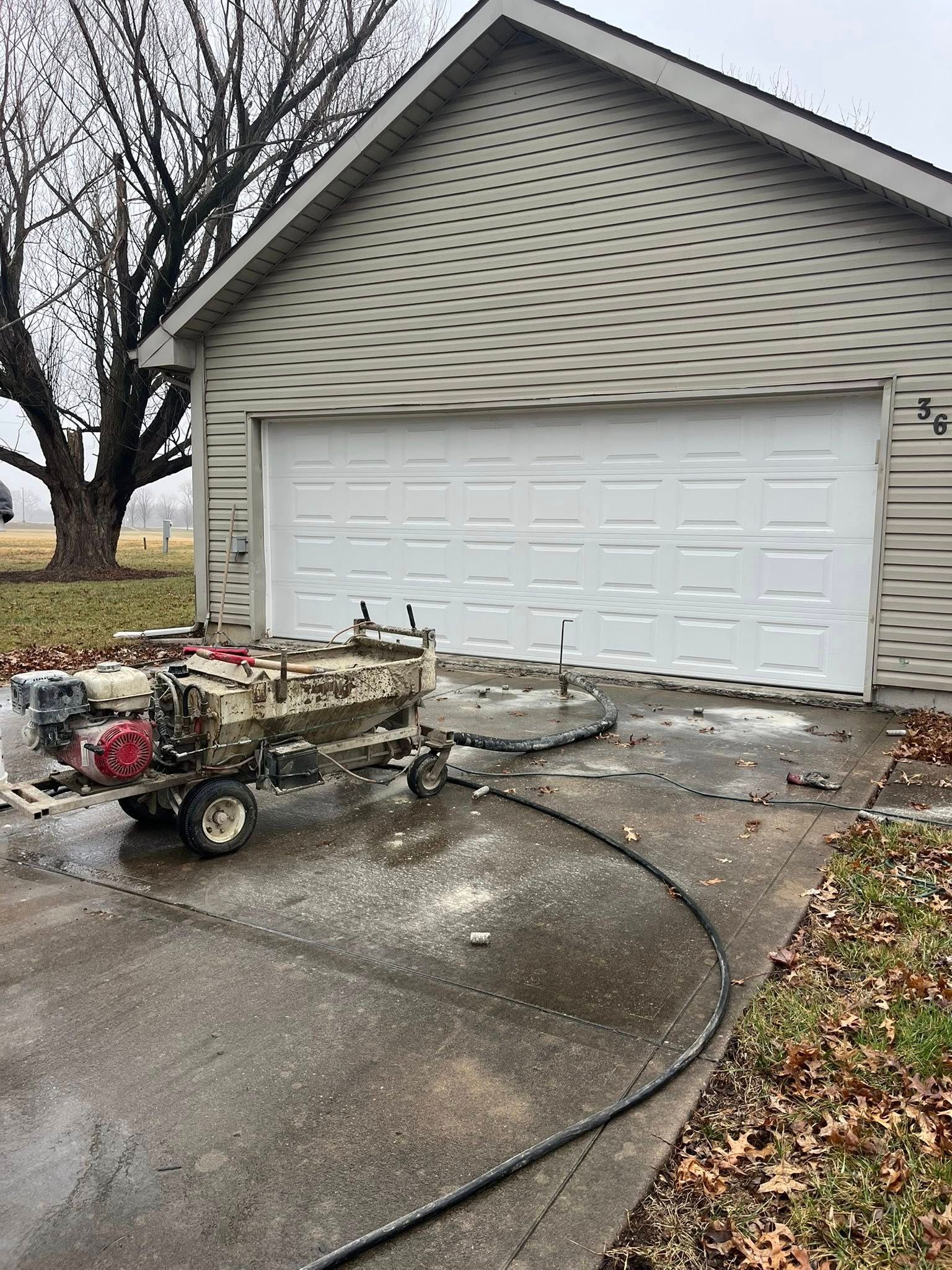 Snow-covered driveway with a small utility cart in front of a garage, yard and trees in winter.