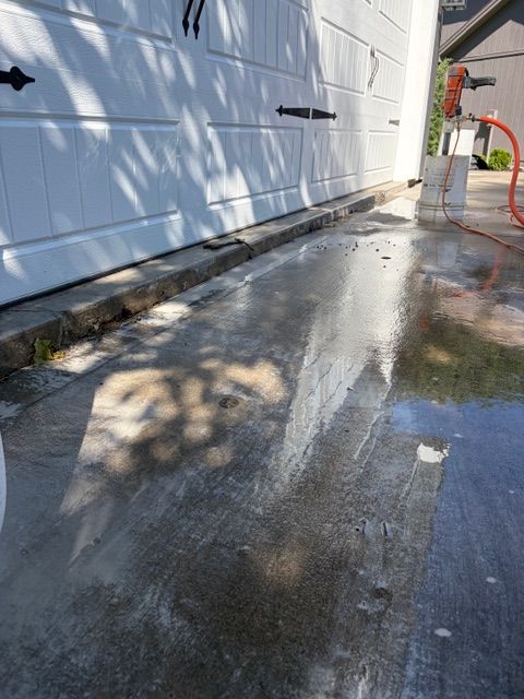 Wet driveway beside white garage wall with water spraying from a hose, creating a reflective surface.