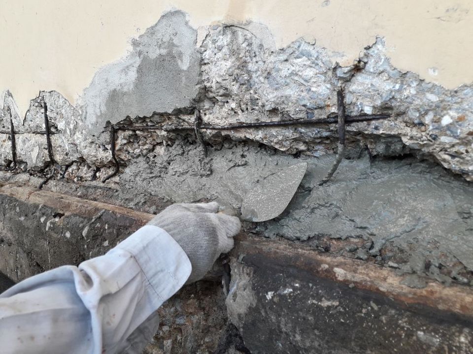 Hand trowel applying gray mortar to a cracked concrete wall near exposed rebar
