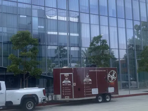 White pickup towing a maroon mobile trailer parked outside a glass office building.