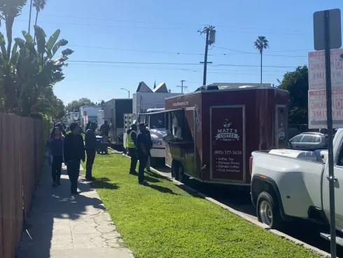 People walking past a red food trailer parked on a sunny residential street